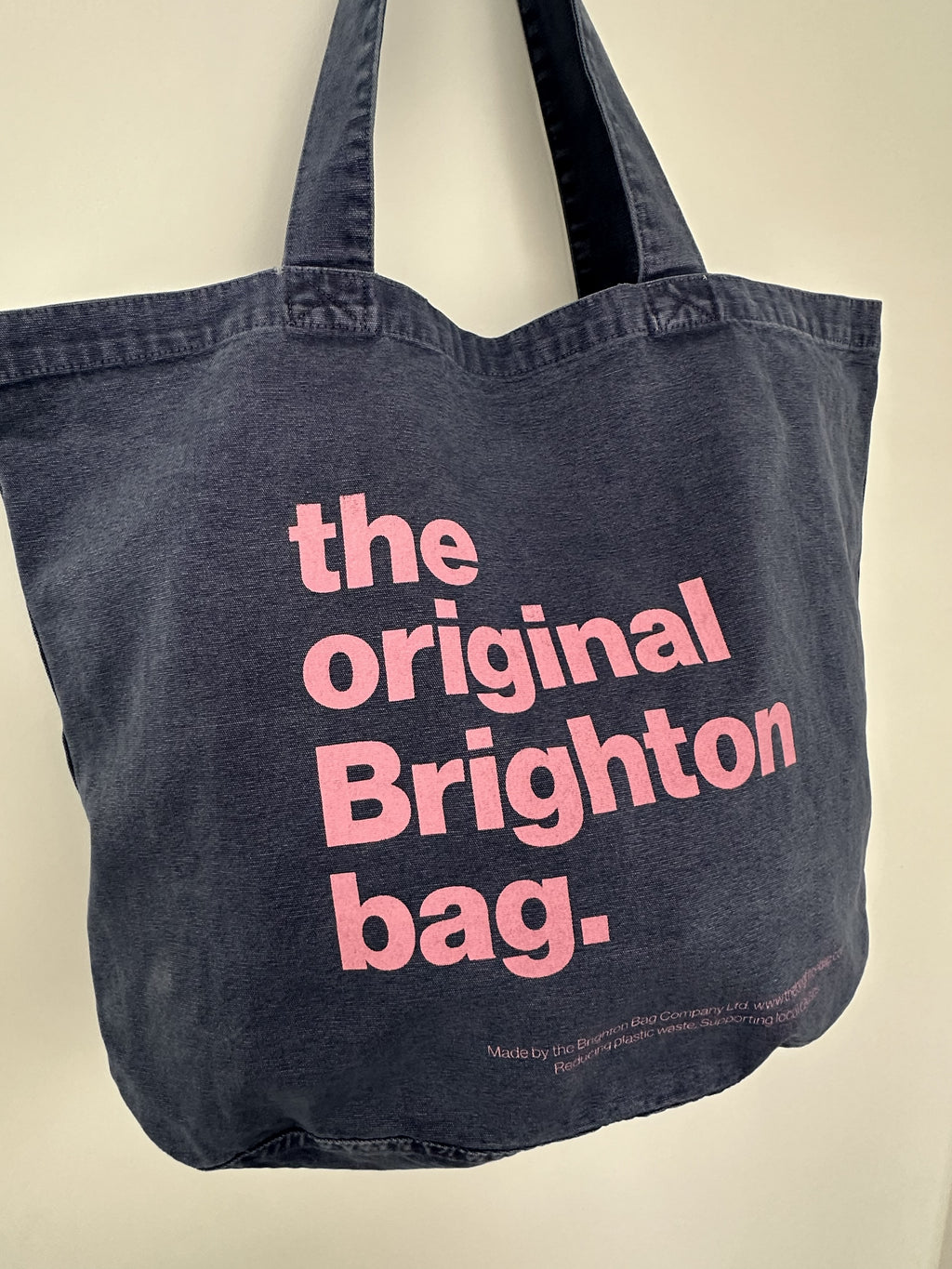 A dark washed denim tote bag with pink text reading 'the original Brighton bag.' hanging against a plain off-white wall.