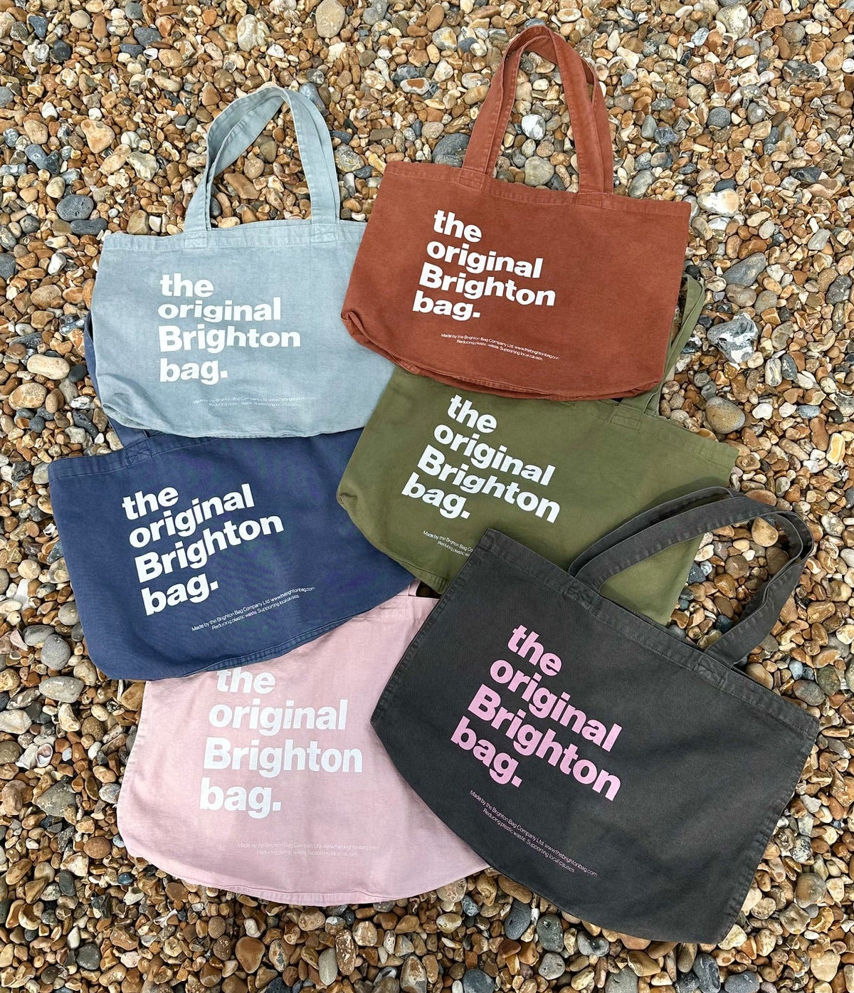 Five tote bags in different colors with 'the original Brighton bag' text on a pebbled surface.