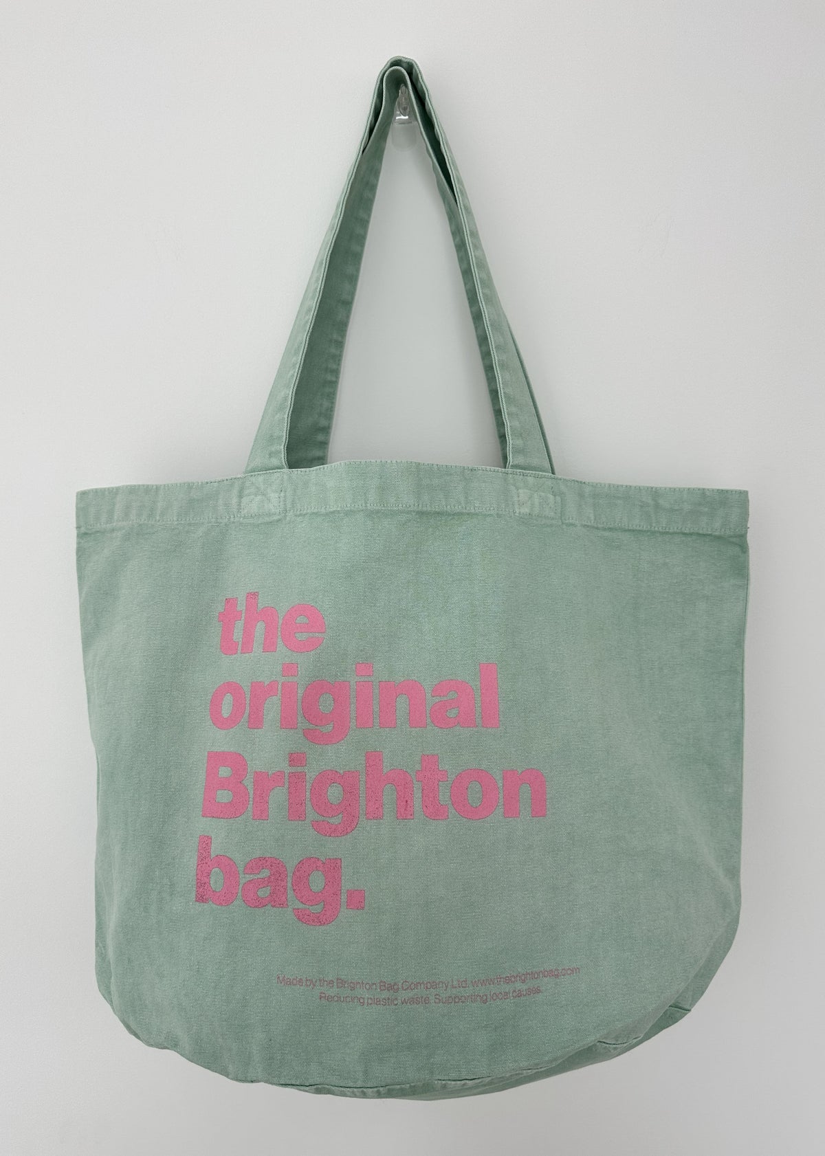 A mint green canvas tote bag with pink text reading 'the original Brighton bag.' printed on the front, hanging against a plain light gray wall.