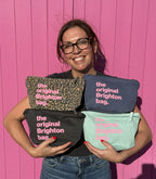 A smiling woman wearing glasses holds four cosmetic pouches in front of a bright pink wooden background. The pouches are in leopard print, navy blue, black, and mint green, each printed with the text 'the original Brighton bag.