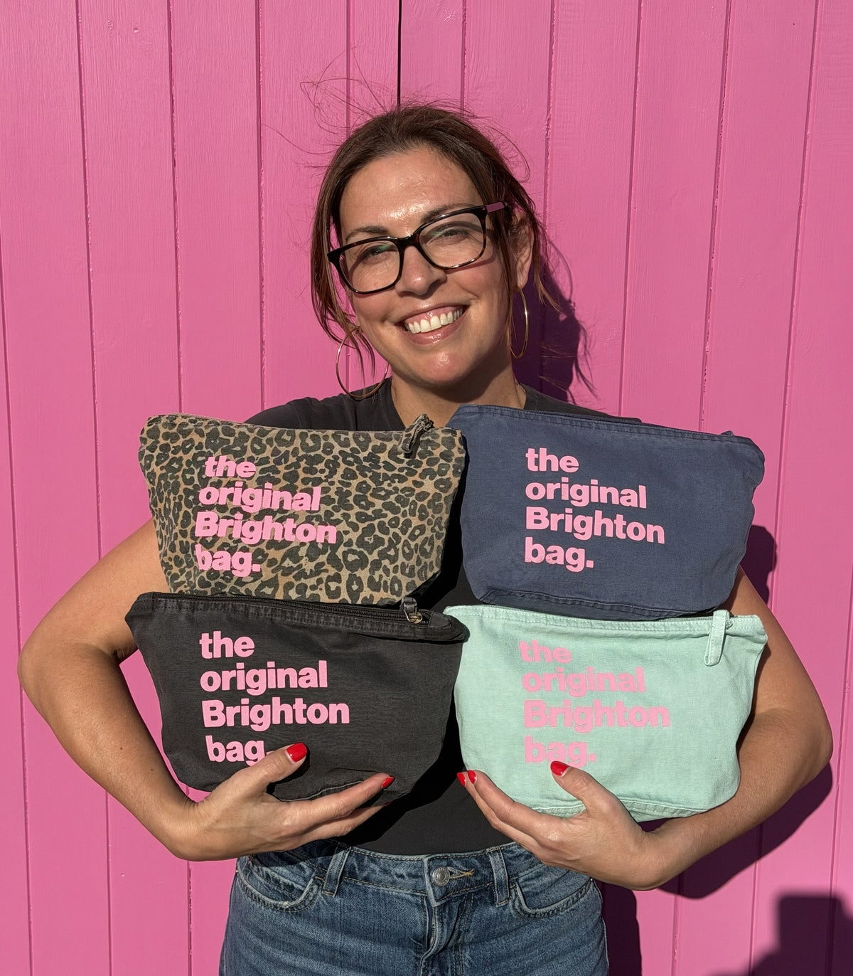 A smiling woman wearing glasses holds four cosmetic pouches in front of a bright pink wooden background. The pouches are in leopard print, navy blue, black, and mint green, each printed with the text 'the original Brighton bag.
