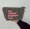 A leopard print cosmetic pouch with pink text reading 'the original Brighton bag.' held by a hand against a plain light background.