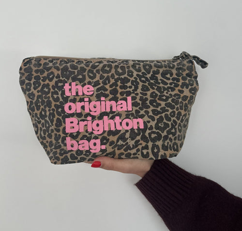 A leopard print cosmetic pouch with pink text reading 'the original Brighton bag.' held by a hand against a plain light background.