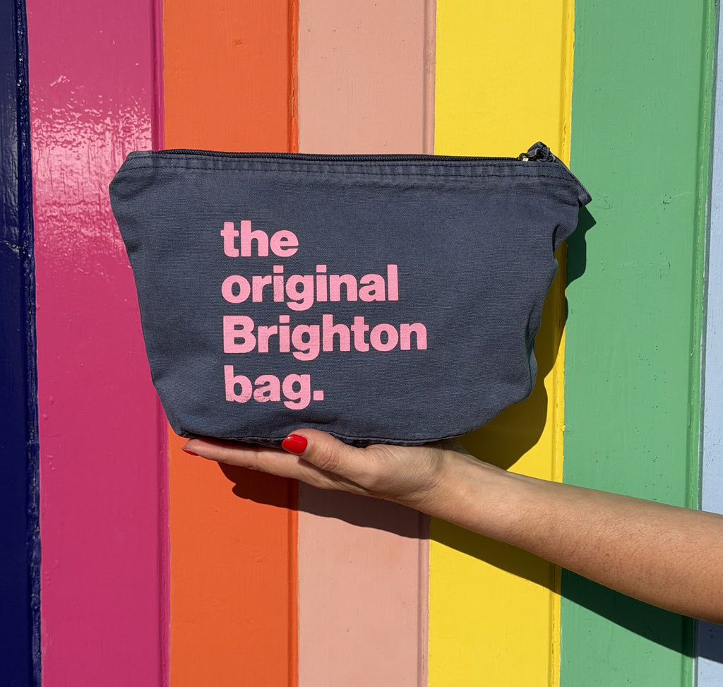 A dark gray fabric pouch with pink text reading 'the original Brighton bag.' is held by a hand against a colorful rainbow-striped background.
