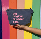 A dark gray fabric pouch with pink text reading 'the original Brighton bag.' is held by a hand against a colorful rainbow-striped background.