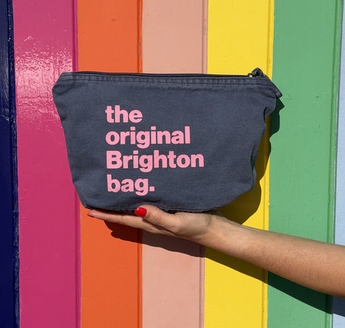 A dark gray fabric pouch with pink text reading 'the original Brighton bag.' is held by a hand against a colorful rainbow-striped background.