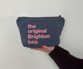 A navy blue fabric zippered pouch with pink text reading 'the original Brighton bag.' held by a hand against a plain light background.