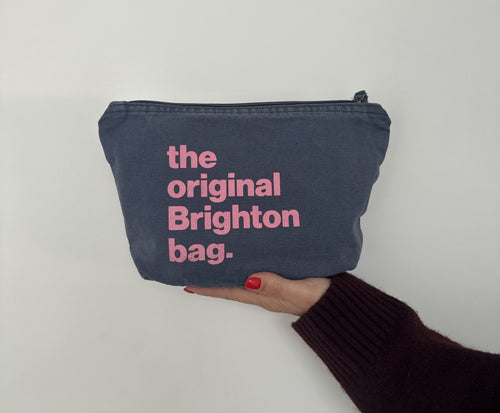 A navy blue fabric zippered pouch with pink text reading 'the original Brighton bag.' held by a hand against a plain light background.