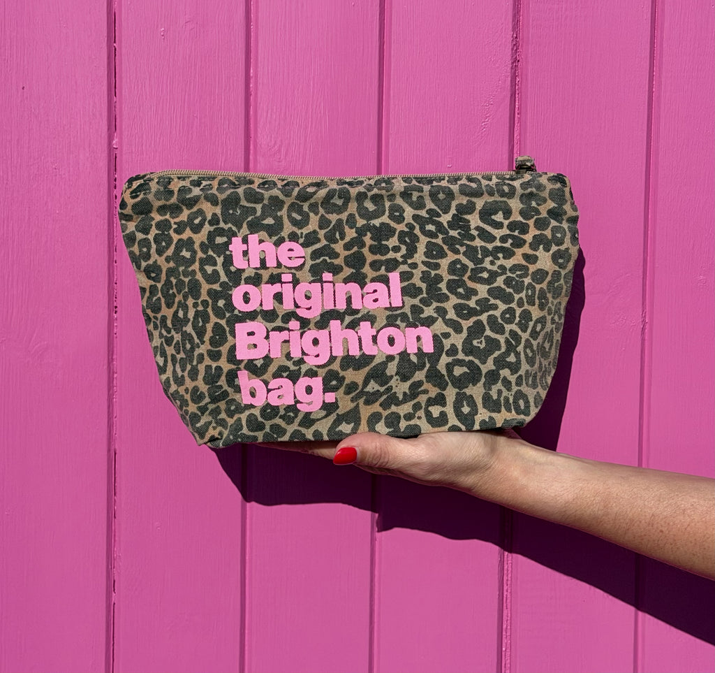 A leopard print cosmetic pouch with pink text reading 'the original Brighton bag.' is held by a hand against a bright pink wooden background.