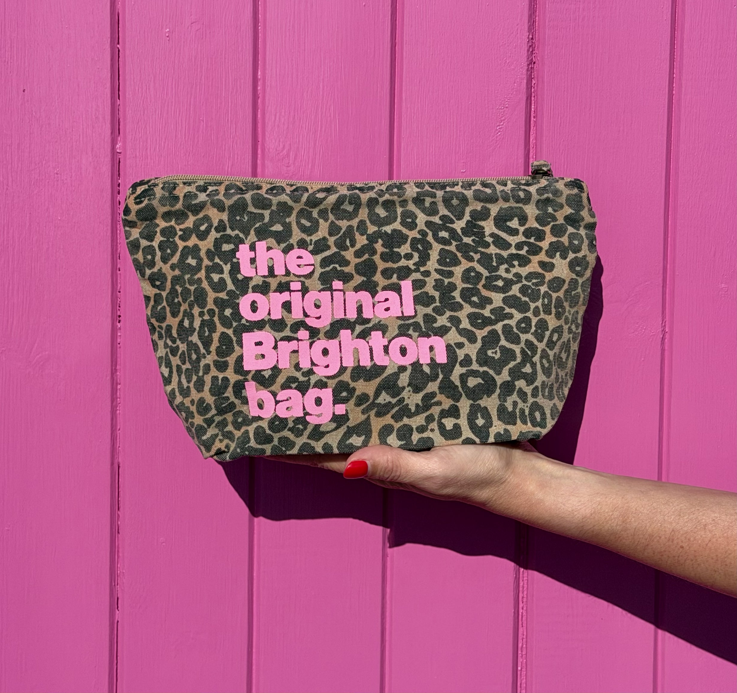 A leopard print cosmetic pouch with pink text reading 'the original Brighton bag.' is held by a hand against a bright pink wooden background.