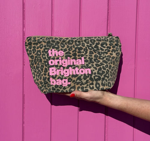 A leopard print cosmetic pouch with pink text reading 'the original Brighton bag.' is held by a hand against a bright pink wooden background.